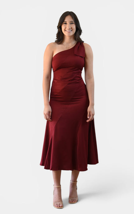 Woman wearing a cranberry one shoulder midi dress with statement bow, fitted bodice, and flared skirt.