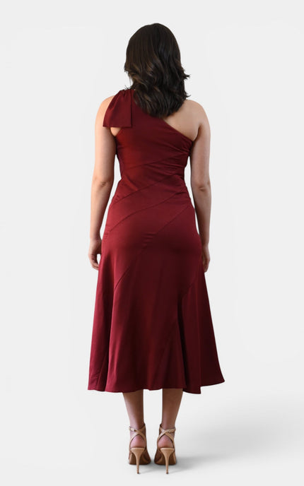 Back view of a cranberry one shoulder midi dress with statement bow shoulder, smooth fitted bodice, and flared midi skirt.