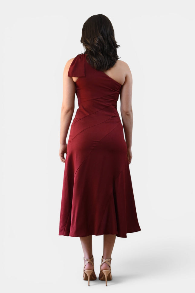 Back view of a cranberry one shoulder midi dress with statement bow shoulder, smooth fitted bodice, and flared midi skirt.