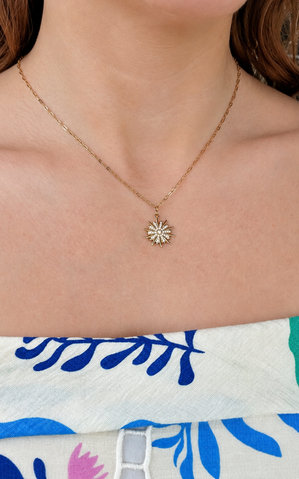 A close-up of a model wearing a delicate gold chain necklace with a small sunburst flower pendant, styled with a blue and white floral off-the-shoulder top.