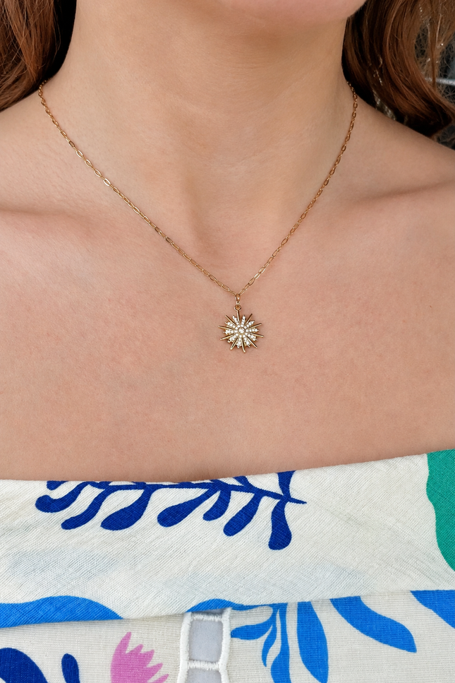 A close-up of a model wearing a delicate gold chain necklace with a small sunburst flower pendant, styled with a blue and white floral off-the-shoulder top.