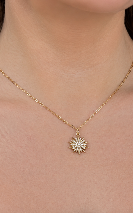 A close-up of a model wearing a delicate gold chain necklace with a small sunburst star pendant, paired with a white top featuring a blue leaf pattern.