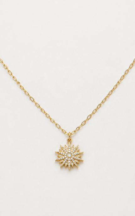 A delicate gold chain necklace with a small sunburst pendant paved with clear crystals, displayed on a clean white background.