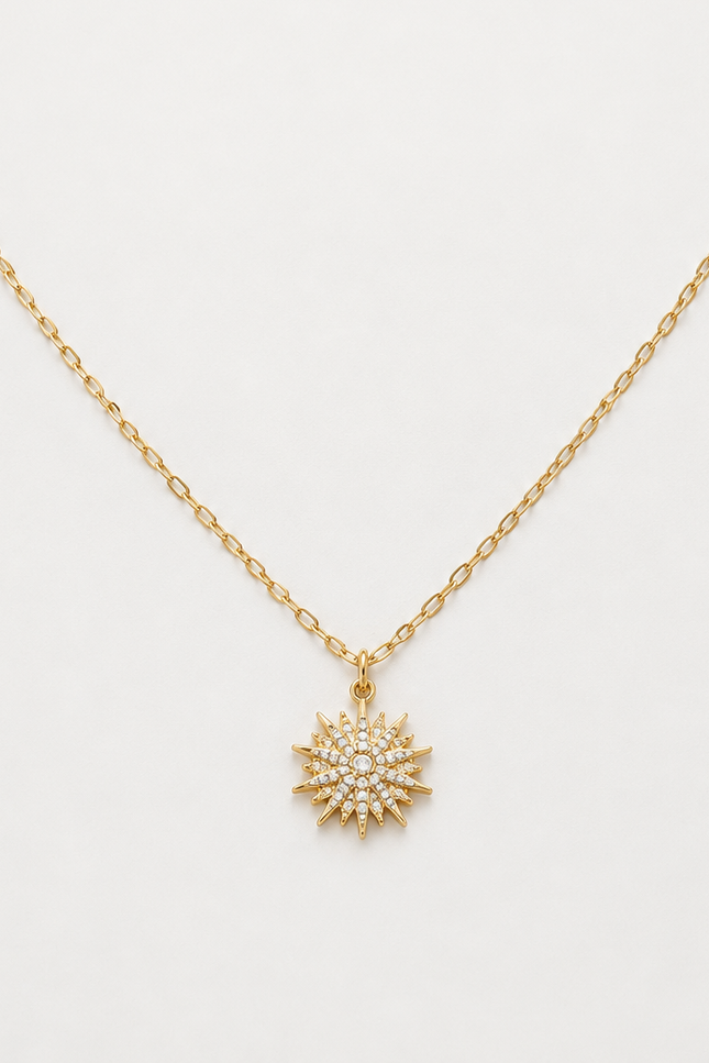 A delicate gold chain necklace with a small sunburst pendant paved with clear crystals, displayed on a clean white background.