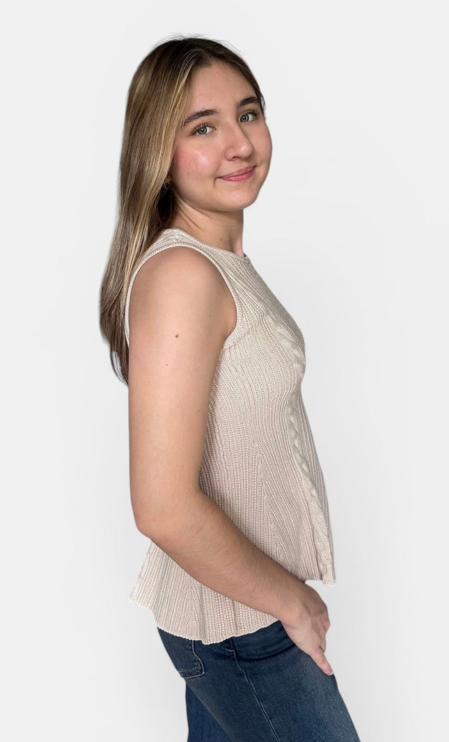 A young woman with blonde-highlighted hair is shown in profile wearing a sleeveless ribbed knit peplum top in a light beige/cream color, paired with blue jeans, against a plain light gray studio background.