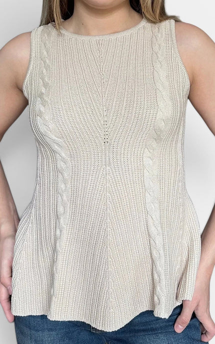 A woman wearing a sleeveless ribbed knit peplum top in a light cream or off-white color, paired with blue jeans, standing against a plain light gray background.