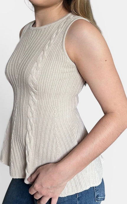A woman modeling a sleeveless ribbed peplum knit top in a light beige color, paired with blue jeans, against a plain light gray background.