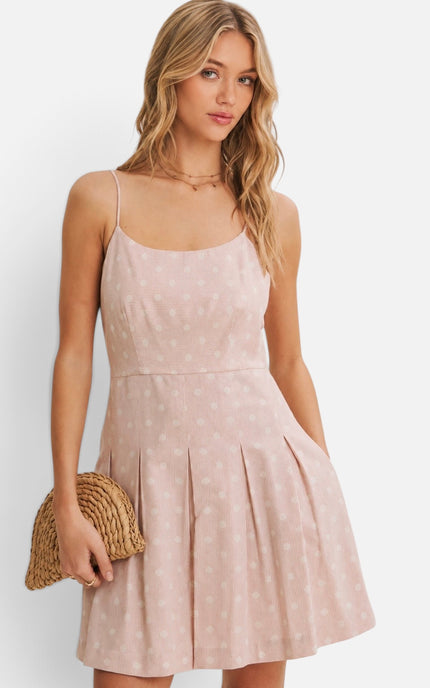 A woman modeling a light pink spaghetti-strap mini dress with a subtle white polka dot print, holding a woven straw clutch against a clean white background.