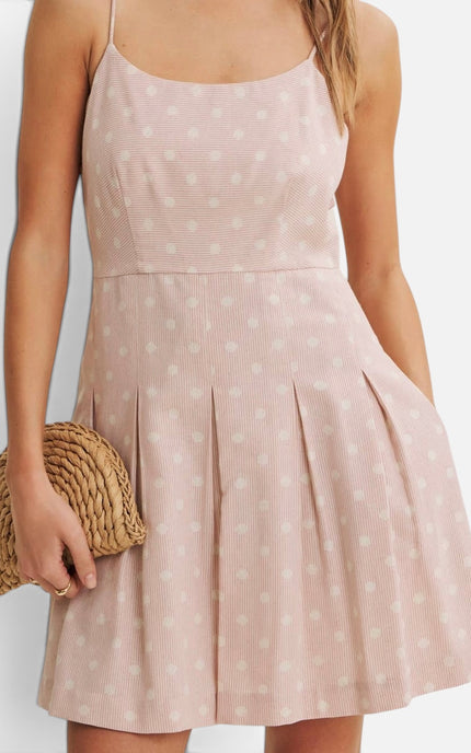 A woman wearing a light pink sleeveless mini dress with white polka dots and a pleated skirt, holding a woven straw clutch against a plain light background.