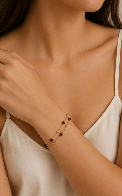 A close-up of a woman wearing a delicate gold chain bracelet with small dark red beads, styled with a white camisole top.