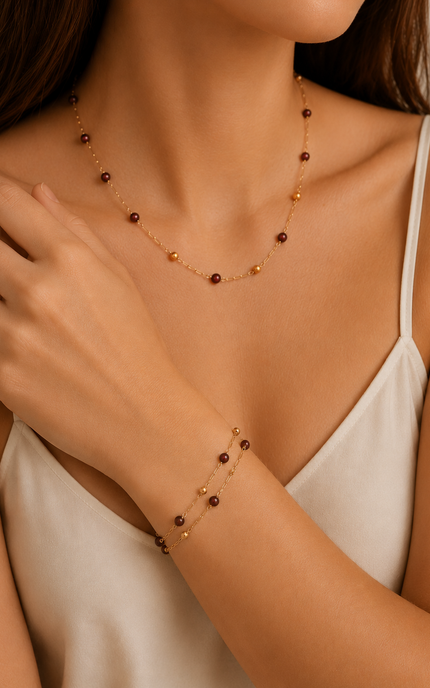 A close-up of a woman wearing a delicate gold necklace and matching bracelet with small dark brown beads, styled with a white camisole.