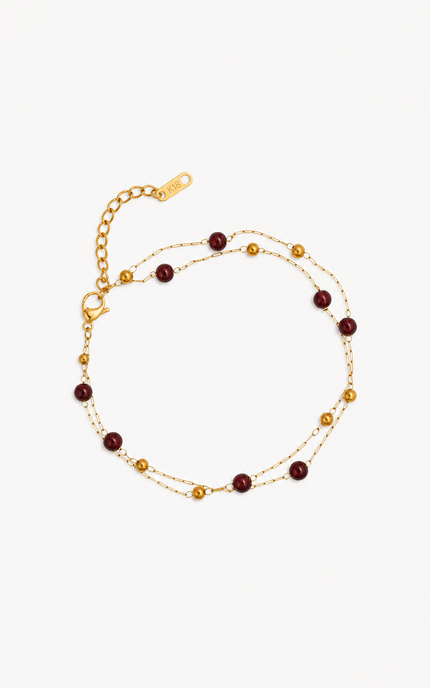 A delicate gold chain anklet featuring alternating small gold beads and round dark red beads, displayed on a clean off-white background.