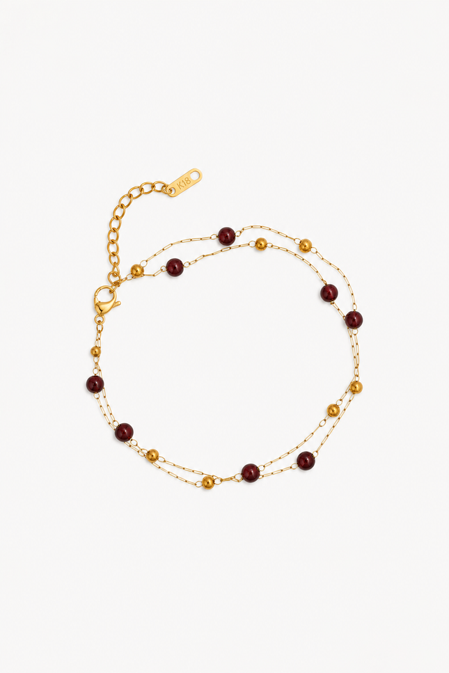A delicate gold chain anklet featuring alternating small gold beads and round dark red beads, displayed on a clean off-white background.