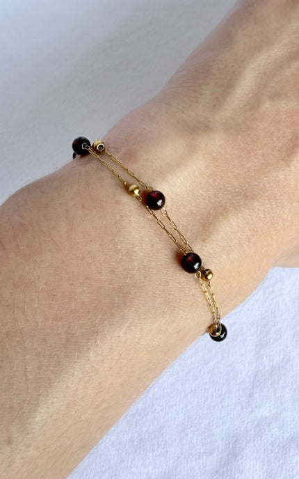 A delicate gold chain bracelet with dark red gemstone beads, shown worn on a person's wrist against a white fabric background.