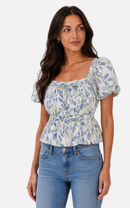 A woman modeling a short-sleeve puff-sleeve top with a blue and green floral print on a cream background, paired with blue jeans, against a light gray studio background.