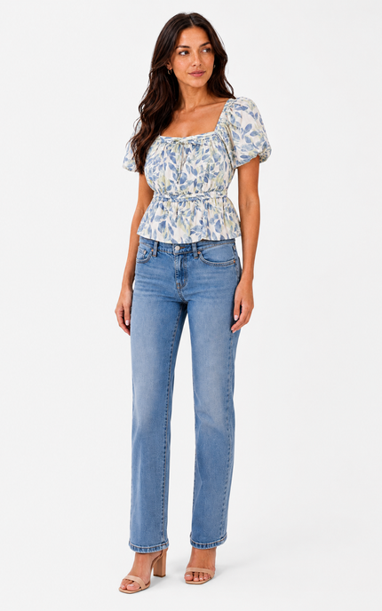 A woman modeling a short-sleeve puff-sleeve peplum top with a blue and white floral print, paired with light-wash blue jeans and nude heeled sandals, against a plain light gray studio background.