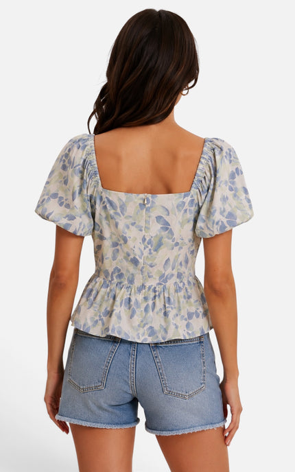A woman shown from the back wearing a short-sleeve peplum top with a blue and gray floral pattern on a cream background, paired with light-wash denim shorts, against a light gray studio background.