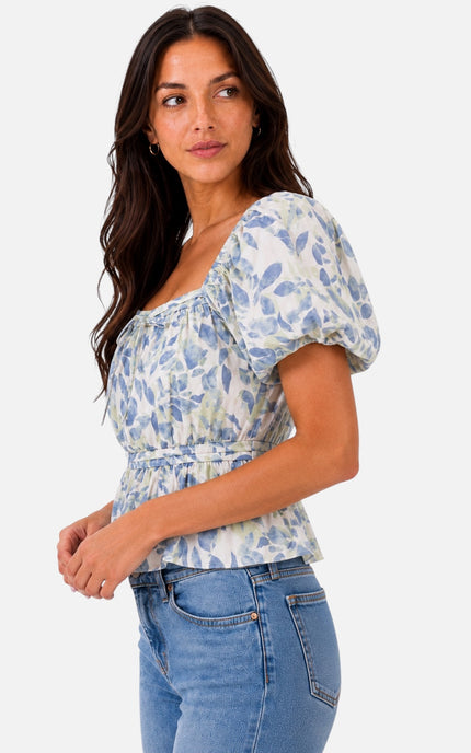 A woman wearing a light blue and white floral print puff-sleeve top and blue jeans, photographed against a plain light gray studio background.