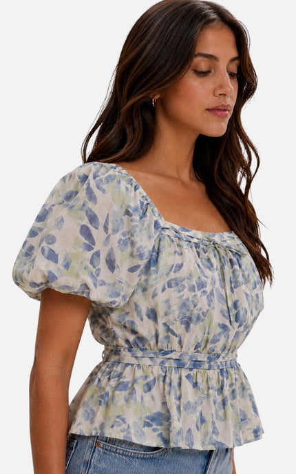 A woman wearing a light blue and white floral print puff-sleeve top with a gathered waist, paired with blue jeans, against a plain light gray background.