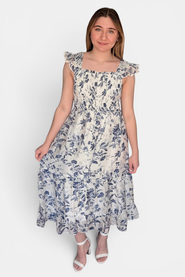 Sleeveless midi dress with a blue floral print on a cream background, featuring ruffled shoulder straps and a smocked bodice, set against a plain light gray studio background.