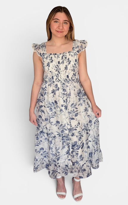 Sleeveless midi dress with a blue floral print on a cream background, featuring ruffled cap sleeves and a smocked bodice, set against a plain light gray background.