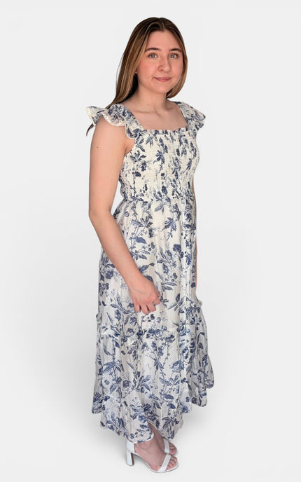 Sleeveless midi dress with a blue floral print on a white background, featuring ruffled shoulder straps and a square neckline, set against a plain light gray studio background.