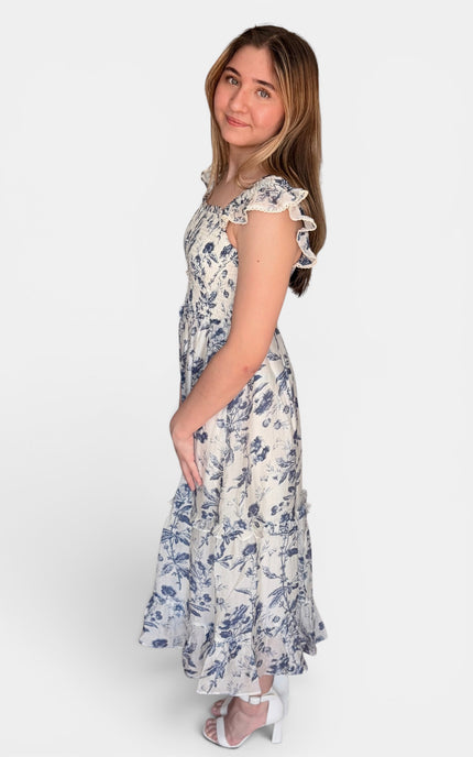 Sleeveless midi dress with a blue floral print on a white background, featuring ruffled shoulder straps and a tiered skirt, shown against a plain light gray studio background.