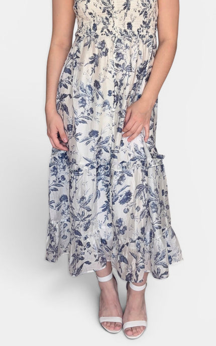 Sleeveless midi dress with a blue floral print on a cream background, featuring ruffled shoulder straps and a tiered skirt, set against a plain light gray studio background.