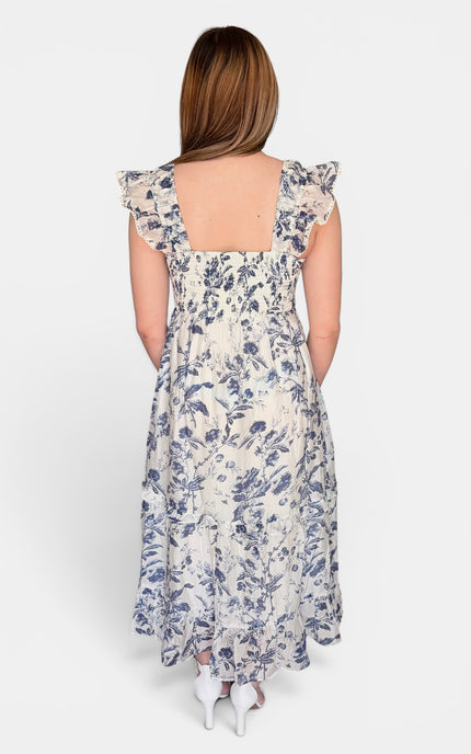 Back view of sleeveless midi dress with a blue floral print on a cream background, featuring ruffled shoulder straps and a tiered skirt, set against a plain light gray studio background.