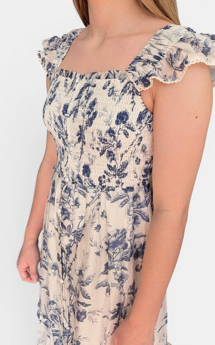Close up of sleeveless midi dress with a blue floral print on a cream background, featuring ruffled shoulder straps and a tiered skirt, set against a plain light gray studio background.