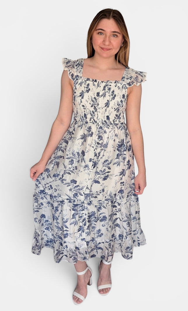 Sleeveless midi dress with a blue floral print on a cream background, featuring ruffled shoulder straps and a smocked bodice, set against a plain light gray studio background.