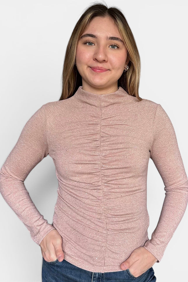 A young woman wearing a long-sleeve, high-neck, ruched knit top in a dusty rose pink color, paired with blue jeans, against a plain light gray background.
