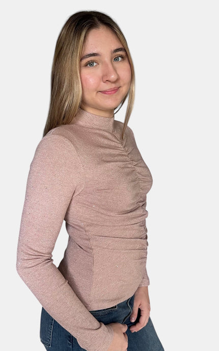 A young woman with blonde hair wearing a dusty rose long-sleeve mock neck top with ruching, paired with blue jeans, photographed against a solid light gray background.