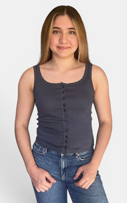 A young woman with long light brown hair wearing a dark gray ribbed tank top with a button-down front and blue jeans, standing against a light gray background.