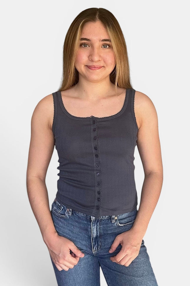 A young woman with long light brown hair wearing a dark gray ribbed tank top with a button-down front and blue jeans, standing against a light gray background.