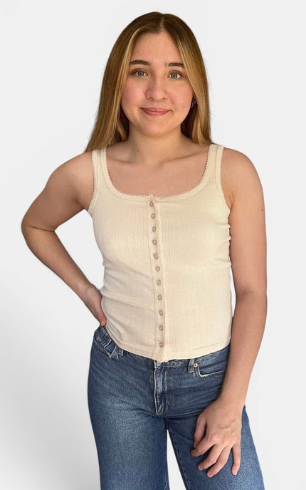 A young woman modeling a cream ribbed tank top with a button-down front and blue jeans, standing against a light gray studio background.