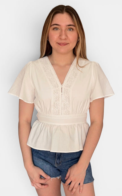 A young woman modeling a cream-colored short-sleeve blouse with a lace V-neckline and peplum hem, paired with denim shorts, against a plain light gray background.