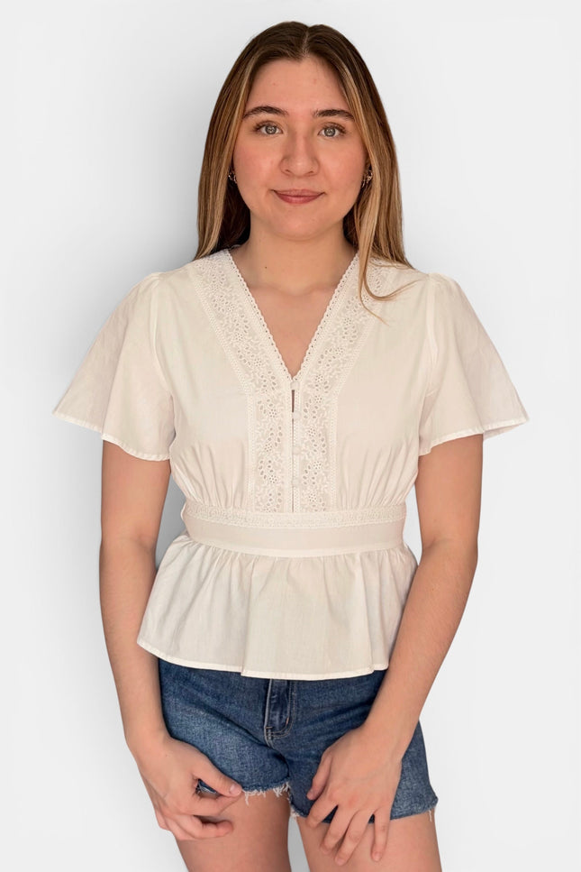A young woman modeling a cream-colored short-sleeve blouse with a lace V-neckline and peplum hem, paired with denim shorts, against a plain light gray background.