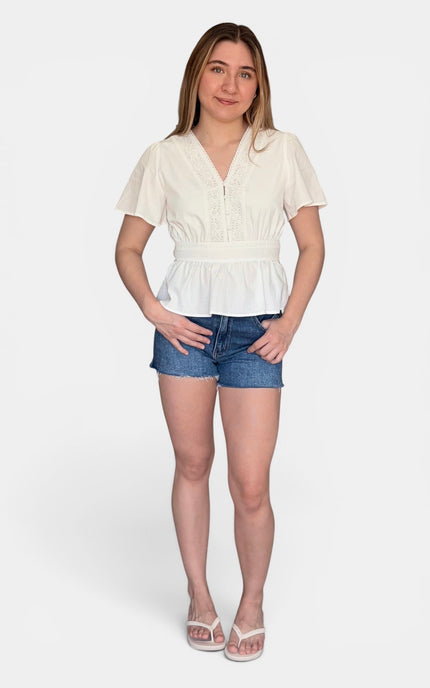 A woman modeling a short-sleeve white peplum blouse with a V-neck and button front, paired with blue denim shorts and white sandals, against a light gray studio background.