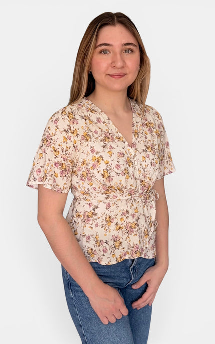 A woman modeling a short-sleeve wrap-style blouse with a beige base and a dense yellow, pink, and green floral print, paired with blue jeans against a plain light gray background.