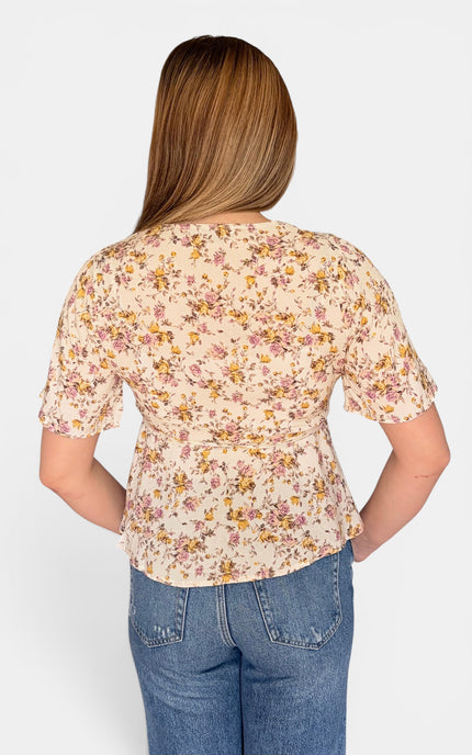 A woman viewed from the back wearing a short-sleeve cream floral top with blue jeans, set against a plain light gray studio background.