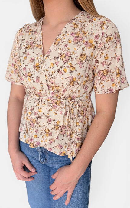 A woman wearing a short-sleeve wrap-style blouse with a beige base and a pink, yellow, and brown floral print, paired with blue jeans against a plain light background.