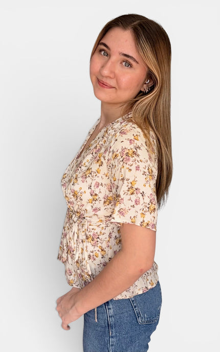 A woman wearing a short-sleeve cream-colored blouse with a yellow and pink floral pattern, paired with blue jeans, photographed against a solid light gray background.
