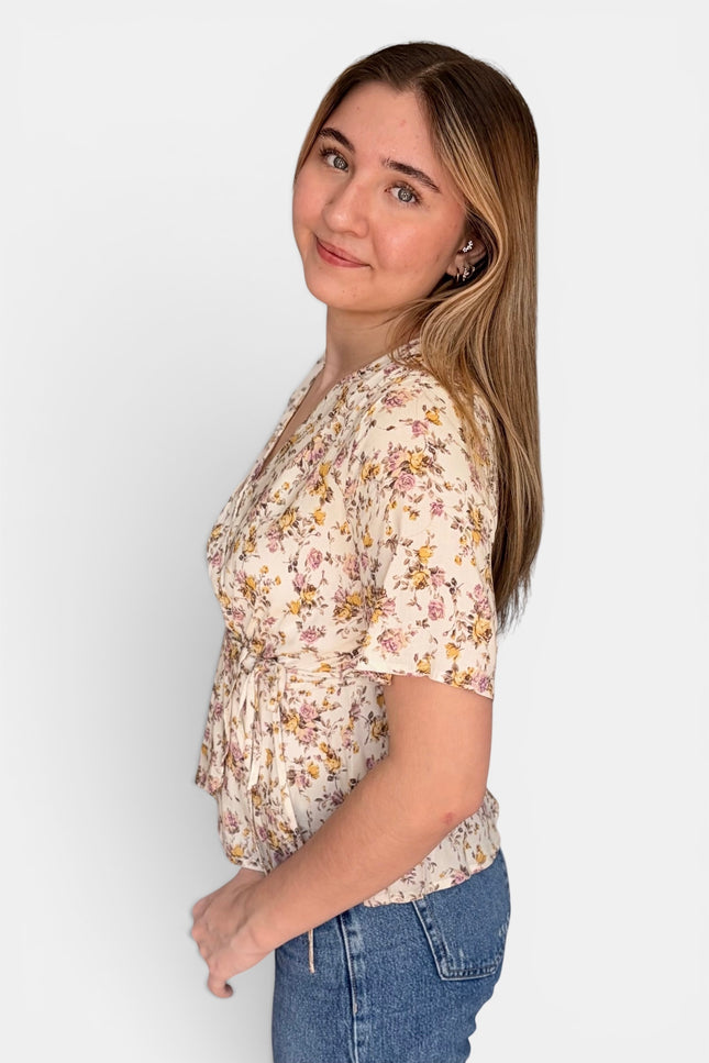 A woman wearing a short-sleeve cream-colored blouse with a yellow and pink floral pattern, paired with blue jeans, photographed against a solid light gray background.