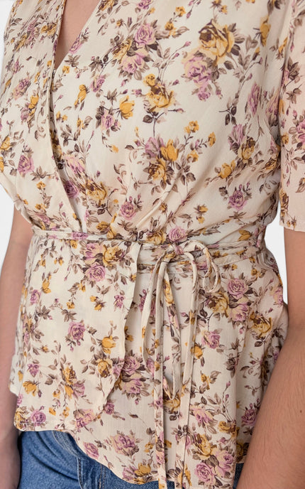 A close-up of a person wearing a cream-colored wrap top with a yellow and pink floral print, tied at the waist, paired with blue jeans.