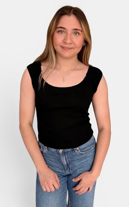 A young woman with blonde hair wearing a black scoop-neck sleeveless top and blue jeans, standing against a light gray background.