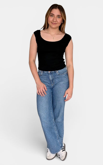 A woman modeling a black scoop-neck sleeveless top and light-wash blue jeans against a plain light gray studio background.