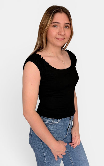 A young woman with light brown hair wearing a black scoop-neck ribbed top and blue jeans, standing against a plain white studio background.