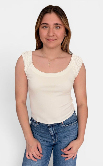 A woman wearing a cream-colored scoop-neck ribbed tank top with ruffled cap sleeves and blue jeans, standing against a plain white background.