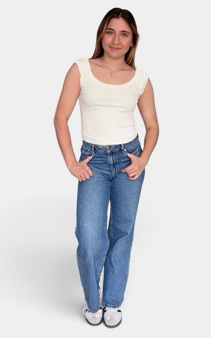 A woman modeling a sleeveless cream top and blue jeans against a light gray studio background.